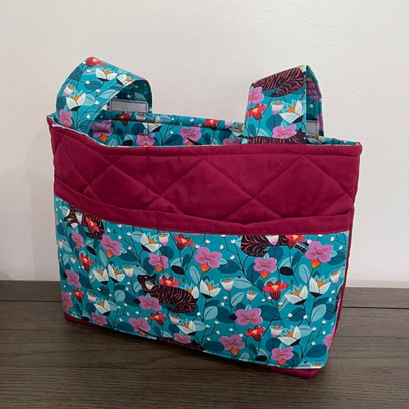 A fabric tote bag with a teal and pink floral print, a solid magenta quilted front pocket, and matching floral shoulder straps, resting on a dark wood surface against a plain white wall.