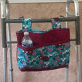 A fabric utility organizer bag hanging from a walker frame. The bag features a teal and pink floral print on the main body and pockets, with a solid burgundy quilted top and bottom trim.