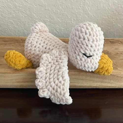 A handmade crocheted plush toy bird, likely a duck or goose, resting on a wooden shelf. The plush is made of chunky off-white yarn with yellow feet and beak, and a small black stitched closed-eye detail.