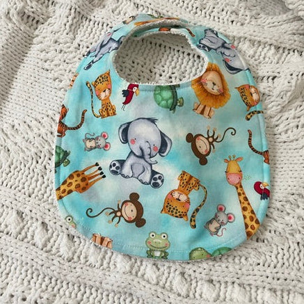 A round baby bib with a light blue background and a colorful safari animal print, laid flat on a textured white knit blanket.