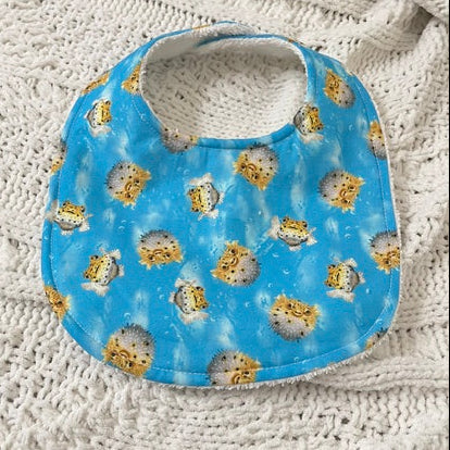 A baby bib with a blue sky and pufferfish pattern, laid flat on a textured cream-colored knit blanket.