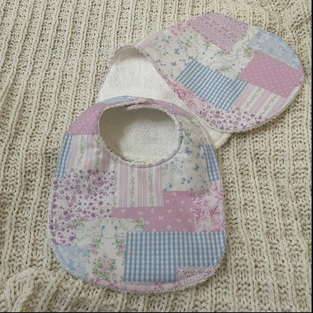 A baby bib with a pastel patchwork print of pink, blue, and white fabrics, displayed on a cream-colored ribbed knit blanket.